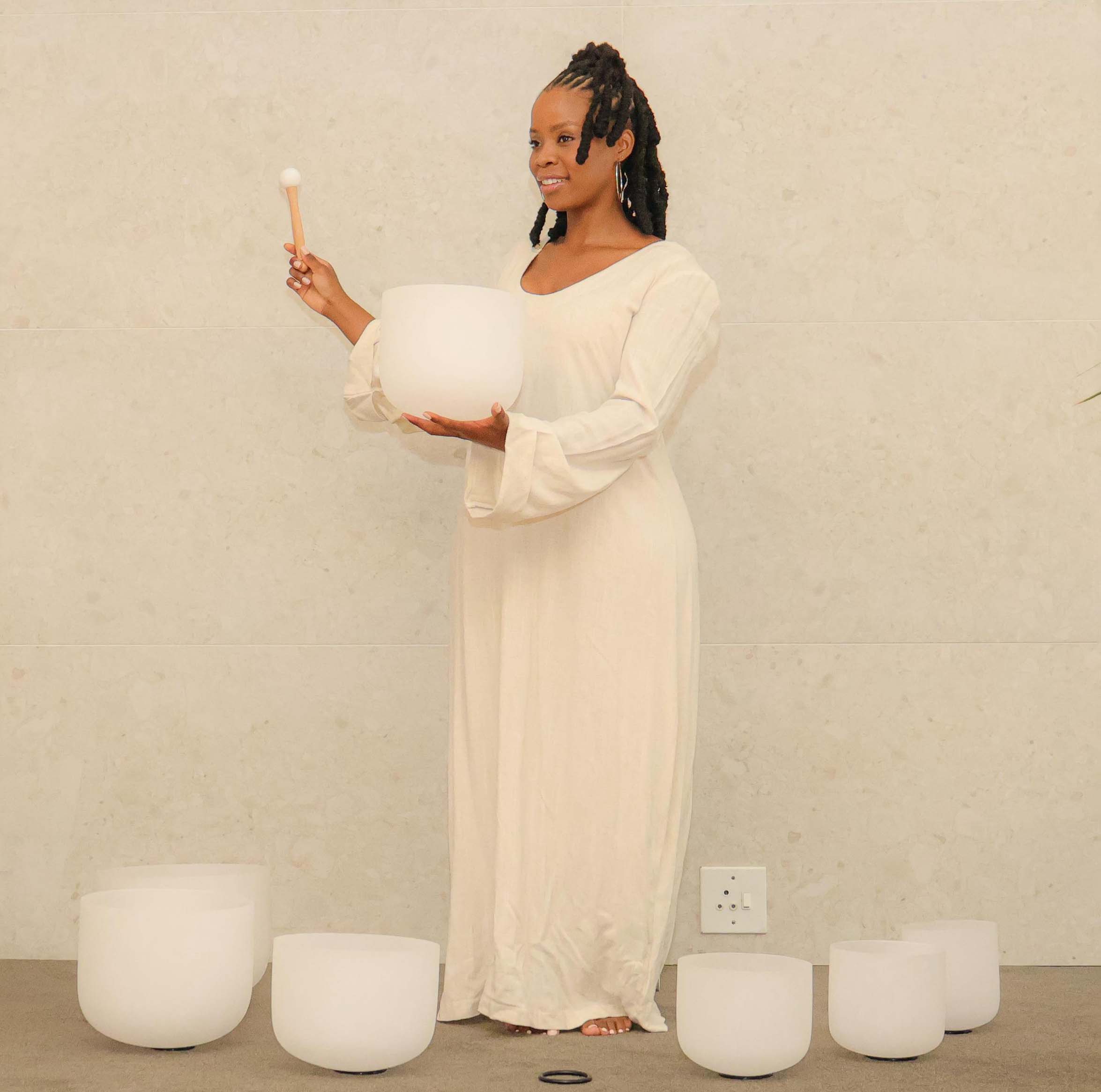 A calming wellness scene featuring a woman in a flowing neutral outfit holding a crystal singing bowl and mallet, surrounded by multiple sound healing bowls. The setting reflects Mint Sanctuary’s holistic approach to healing, using sound therapy to restore balance, reduce stress, and promote inner peace.